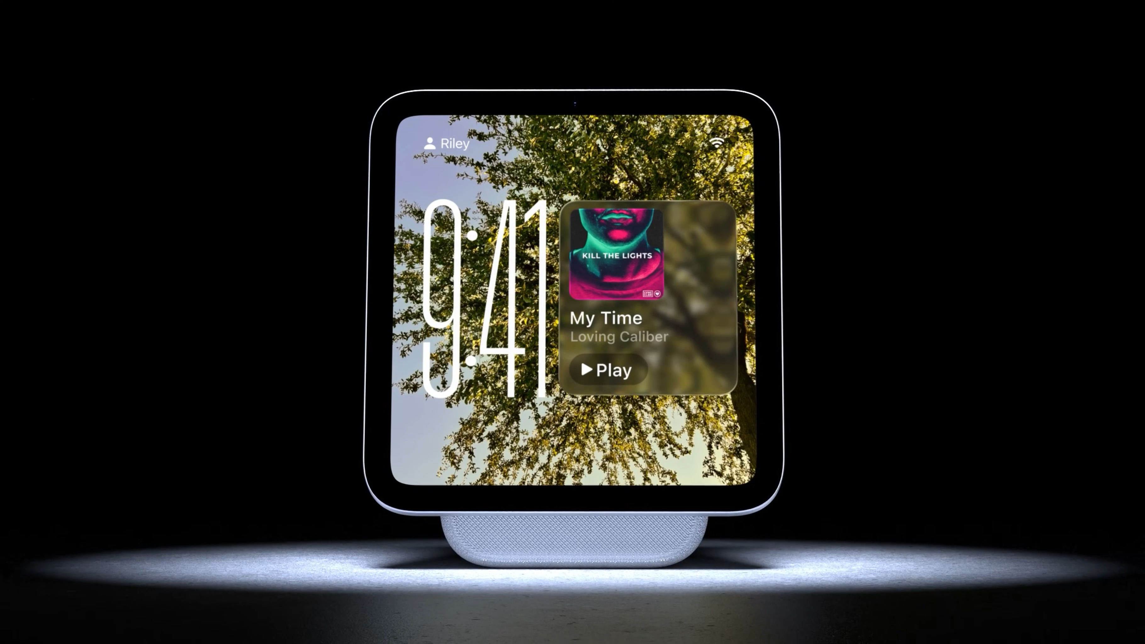Auto-generated description: A smart home device displays a large digital clock and music screen against a backdrop of tree branches.
